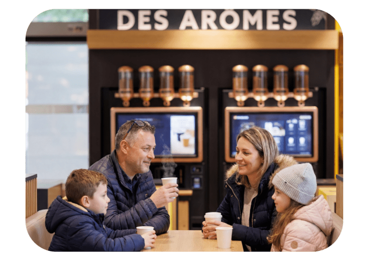 Famille en pause dans une station‑service TotalEnergies, parents dégustant un café offert et enfants un chocolat chaud grâce à l’option Zen de Charge+